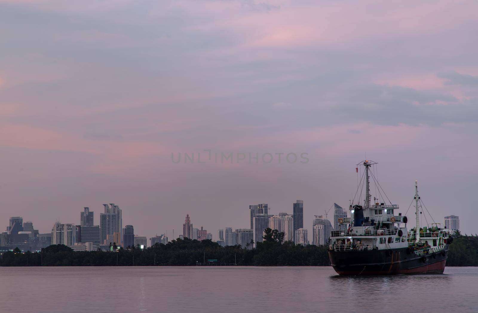 Bangkok, Thailand - 04 Jan 2021 : A cargo ship parked in the middle of the Chao Phraya River at evening with green forest and skyscrapers at the background. Selective focus.