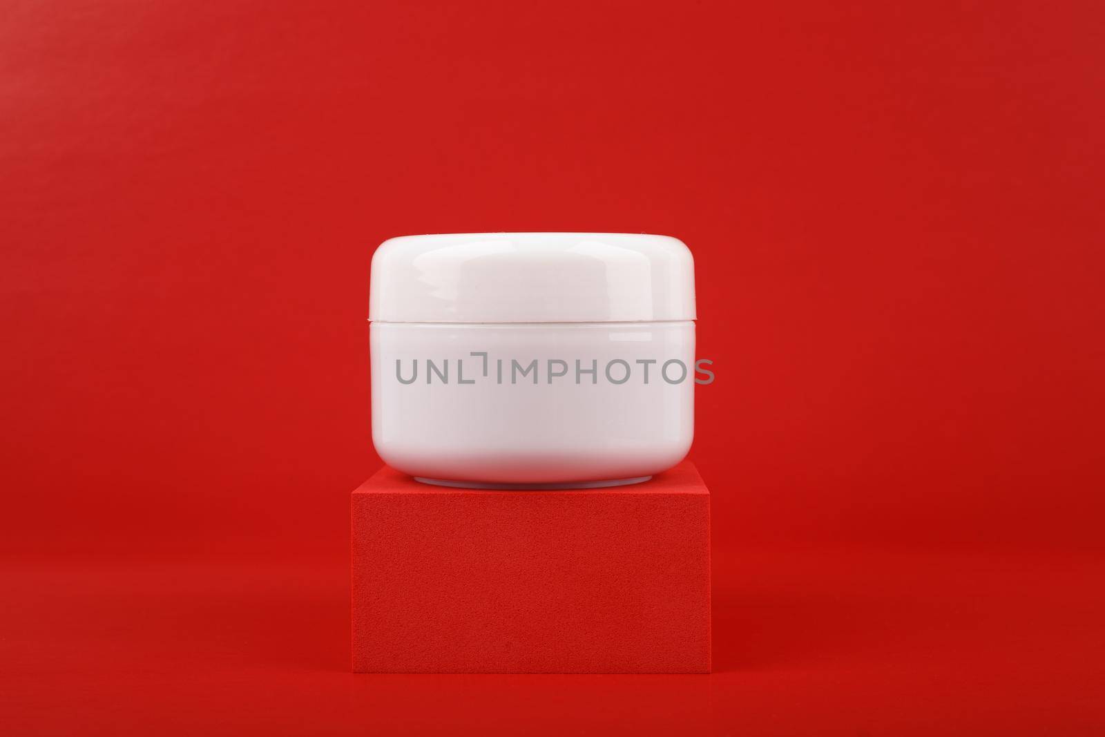 Close up of white glossy cosmetic jar on red podium against red background. 
