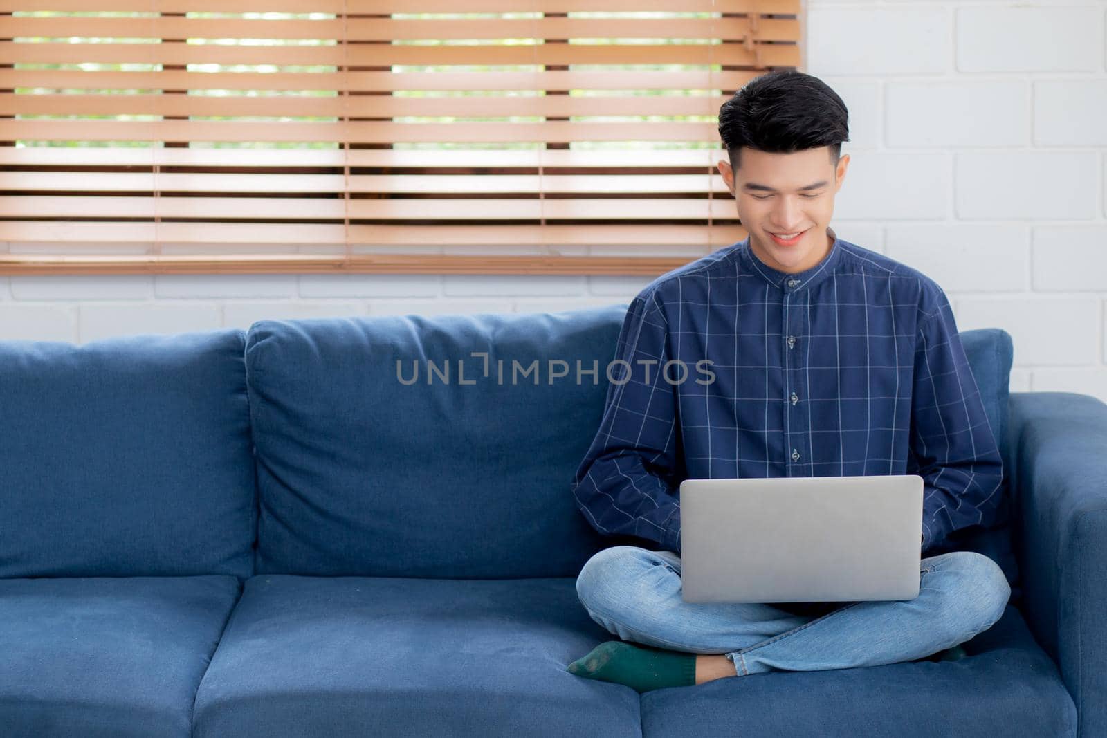 Young asian businessman smile and work from home with laptop computer online to internet on sofa in living room, freelance man using notebook on couch with comfort, new normal, lifestyle concept.