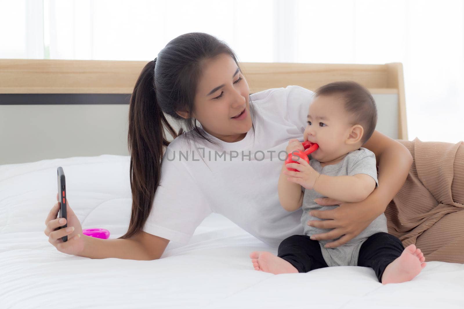 Young asian mother and little baby girl or newborn selfie with smart phone on bed in bedroom, happiness mom and daughter using phone video call at home, two people, family and communication concept. by nnudoo