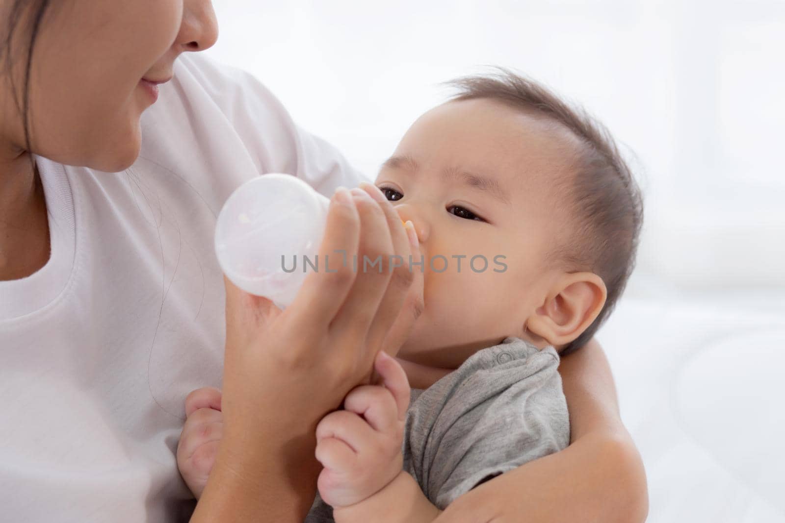 Young asian mother embracing and feeding little baby girl with bottle of milk at home, newborn innocence drinking with mom satisfied, relationship and bonding of mum and child, family concept.