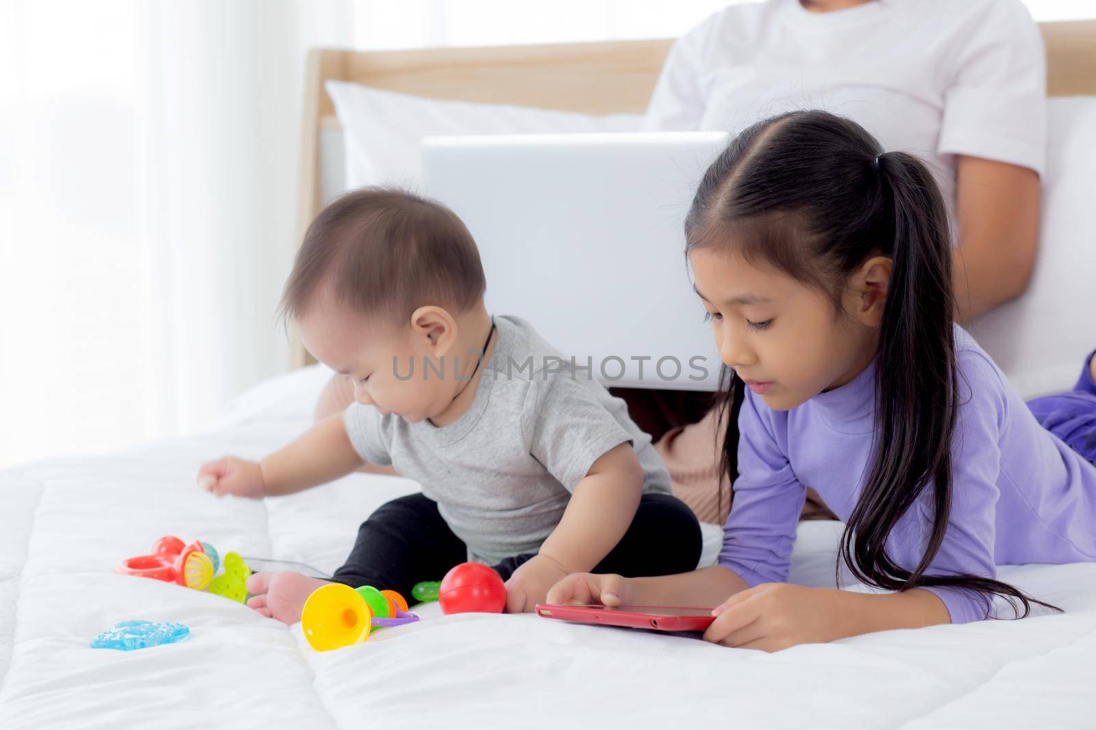 Young asian mother working at home online to internet with laptop computer and daughter looking tablet on bed at bedroom, business woman is freelance and children learning, new normal, family concept. by nnudoo