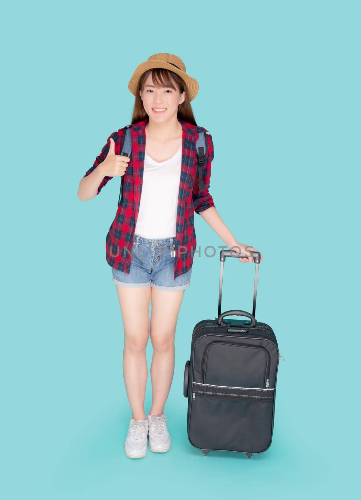 Beautiful young asian woman pulling suitcase isolated on blue background, asia girl cheerful holding luggage walking and gesture thumbs up in vacation with excited, journey and travel concept.
