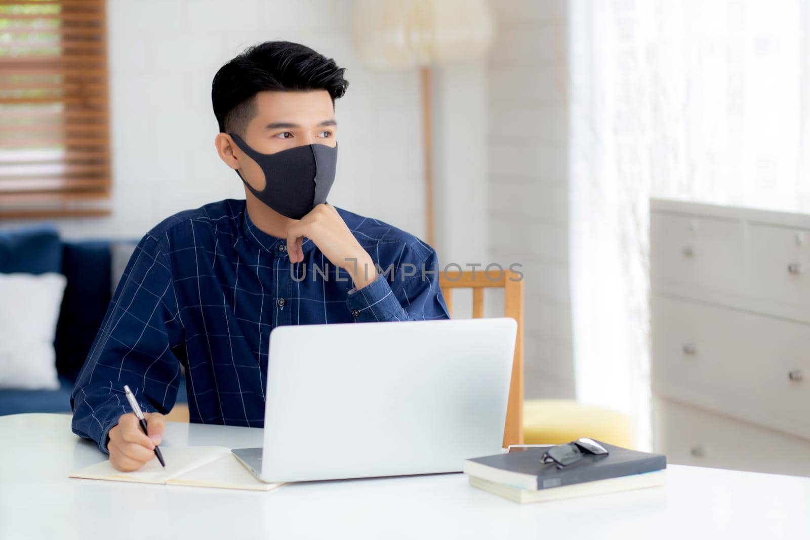 Young business man in face mask planning and working from home on laptop computer on desk for protection for outbreak of covid-19, male quarantine stay home using notebook on desk, new normal.