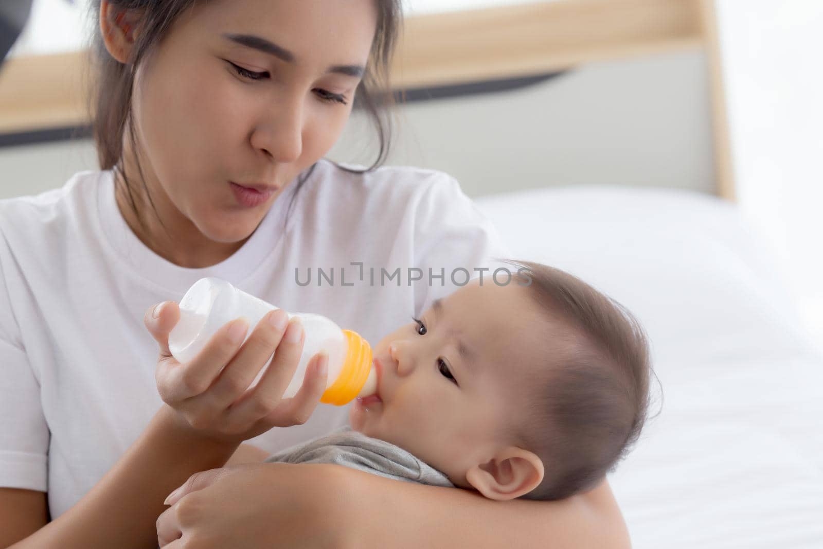 Young asian mother embracing and feeding little baby girl with bottle of milk at home, newborn innocence drinking with mom satisfied, relationship and bonding of mum and child, family concept.