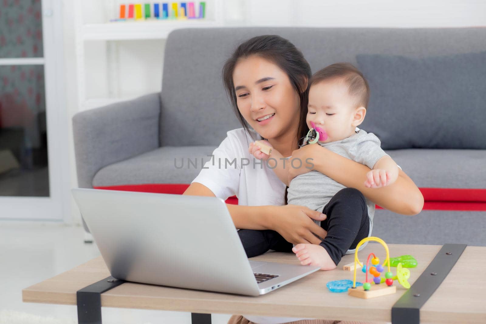 Young asian mother working on laptop computer while care daughter girl at home, mom and baby girl sitting using notebook video call, parent and bonding, indoors, family and business concept.