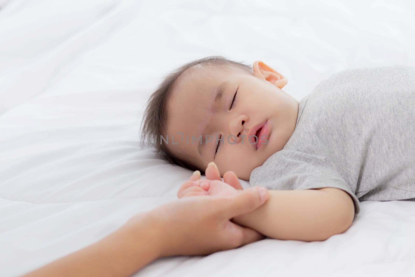 Young asian mother touch hand of little baby girl with tender on bed in the bedroom, mom love newborn and care, woman with expression with child together, parent and daughter, family concept.