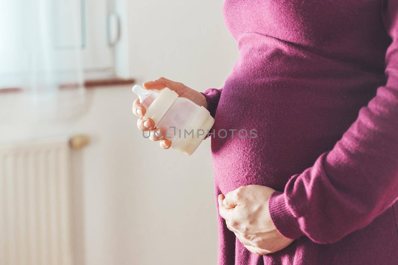 Close up of young mother holding a baby bottle on her pregnant tummy, Caucasian, magenta dress