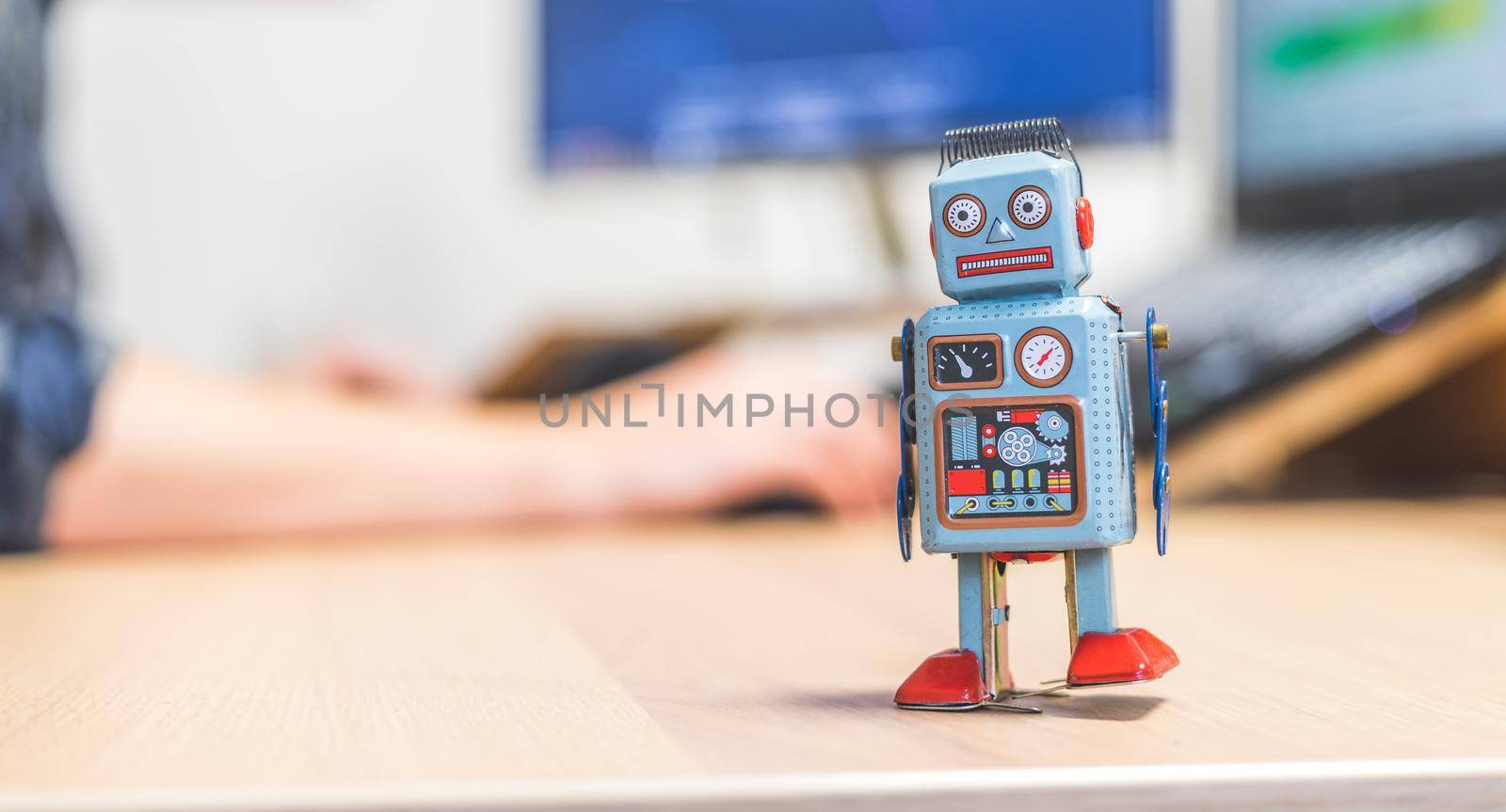 Toy robot on a desktop, man working on the computer in the background. Symbol for artificial intelligence, chatbots or social bots and algorithms