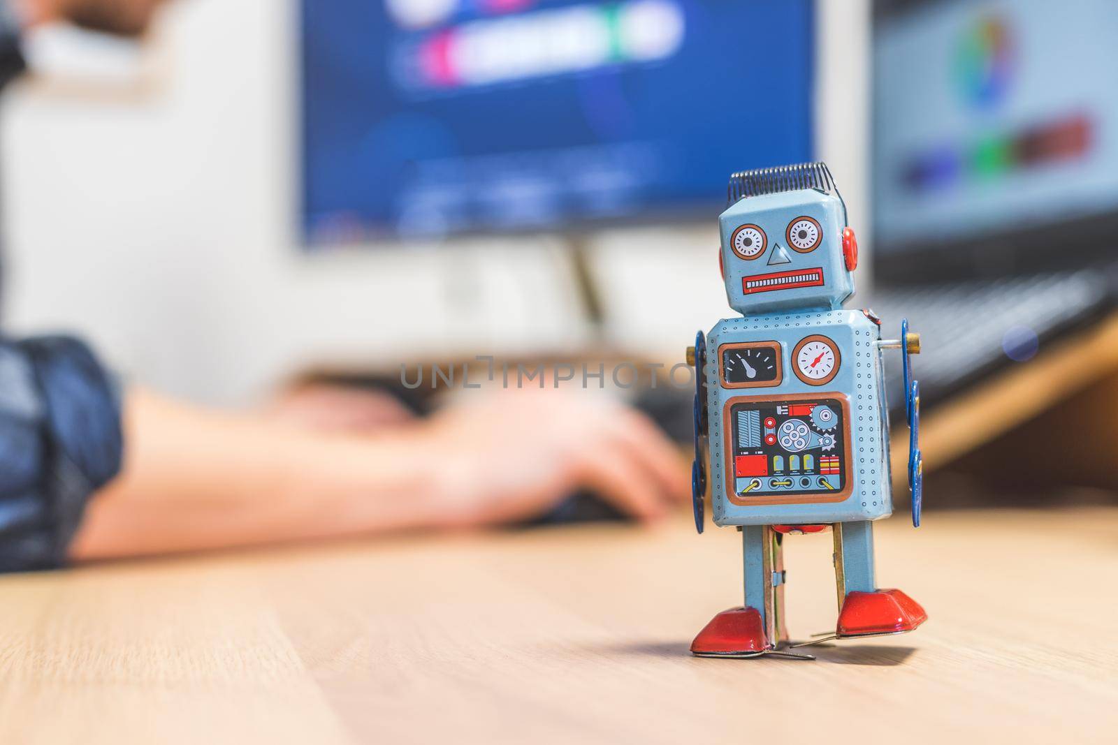 Toy robot on a desktop, man working on the computer in the background. Symbol for artificial intelligence, chatbots or social bots and algorithms
