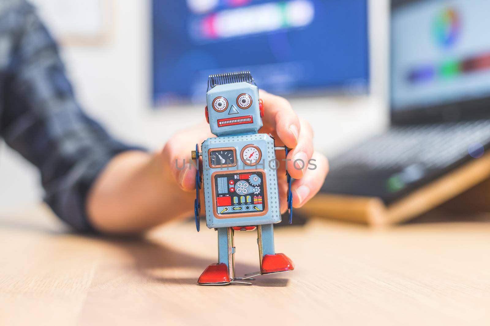 Toy robot on a desktop, man working on the computer in the background. Symbol for artificial intelligence, chatbots or social bots and algorithms