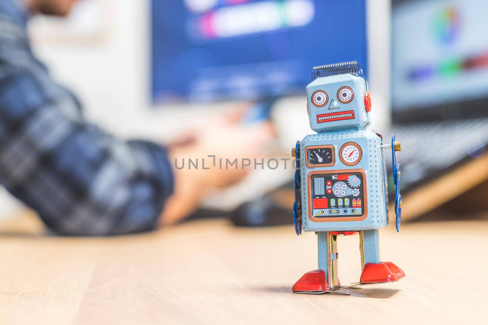 Toy robot on a desktop, man working on the computer in the background. Symbol for artificial intelligence, chatbots or social bots and algorithms
