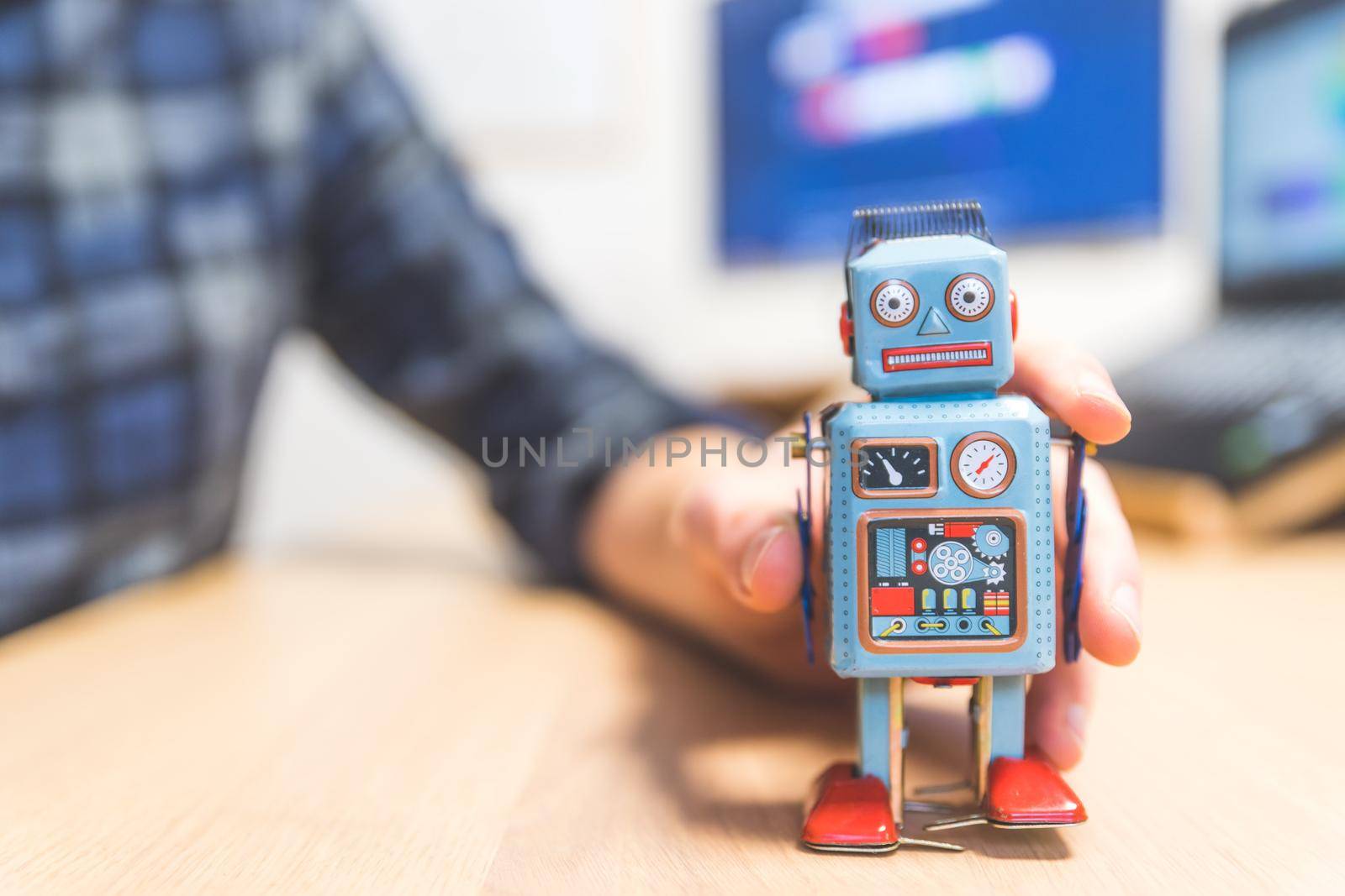 Toy robot on a desktop, man working on the computer in the background. Symbol for artificial intelligence, chatbots or social bots and algorithms