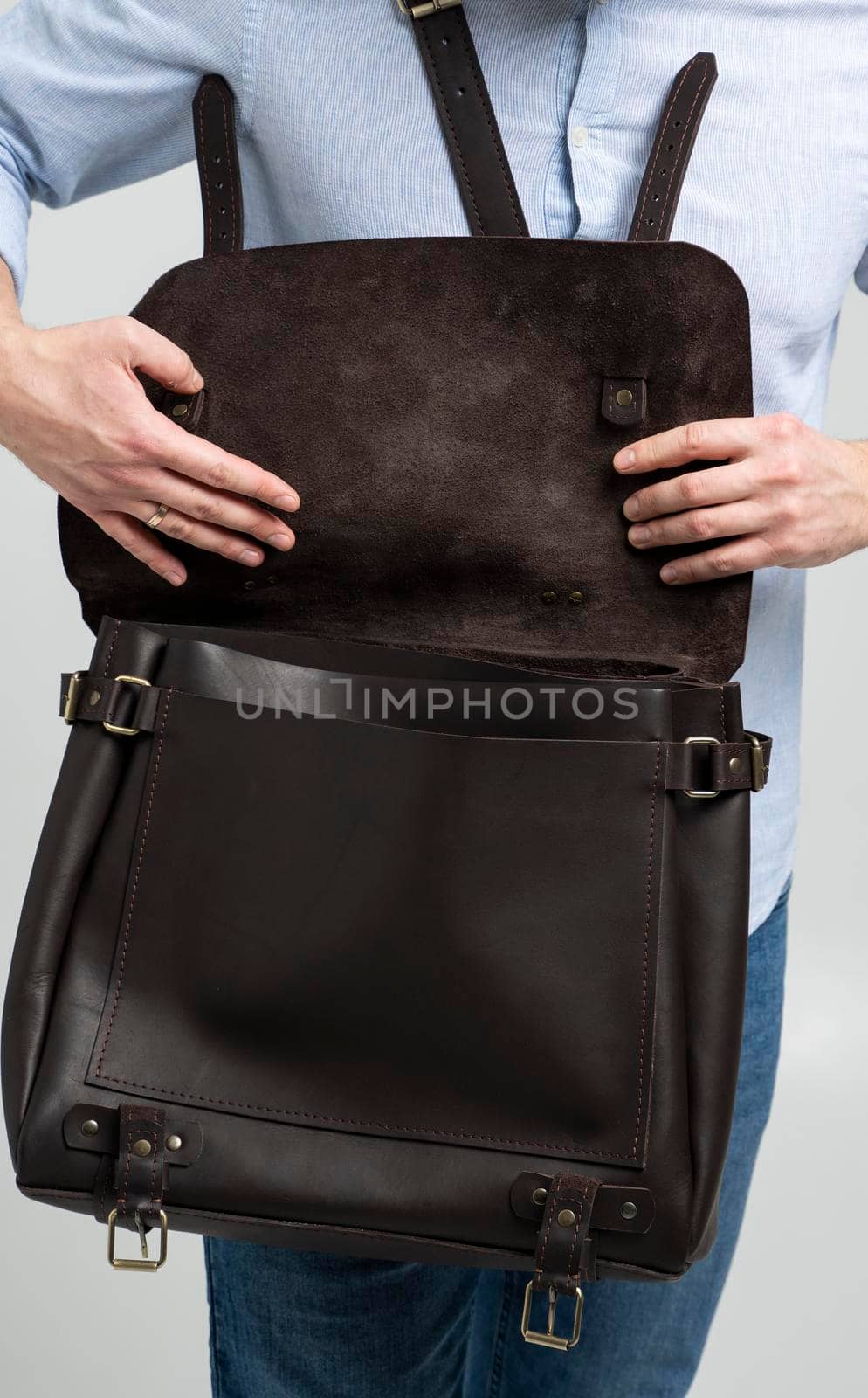 Details of open brown men's shoulder leather bag for a documents and laptop on the shoulders of a man in a blue shirt and jeans with a white background. Satchel, mens leather handmade briefcase