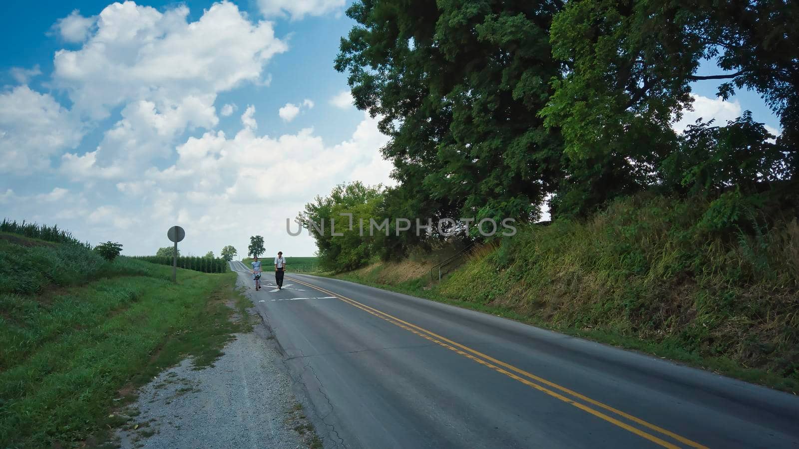 Two Amish Teenagers One Boy and One Girl on Scooters on a Road on a Summer Day