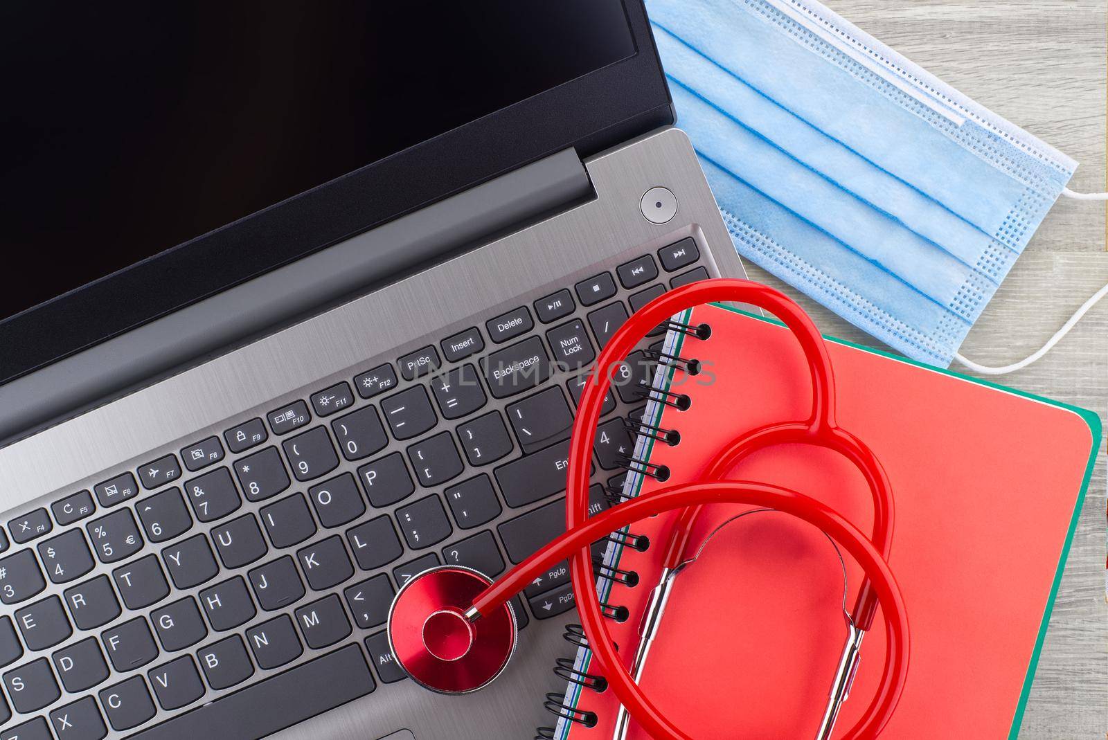 Virus pandemic and medical concept with a colorful red stethoscope coiled over a surgical face mask alongside a notebook on a wooden desk with computer keyboard viewed top down