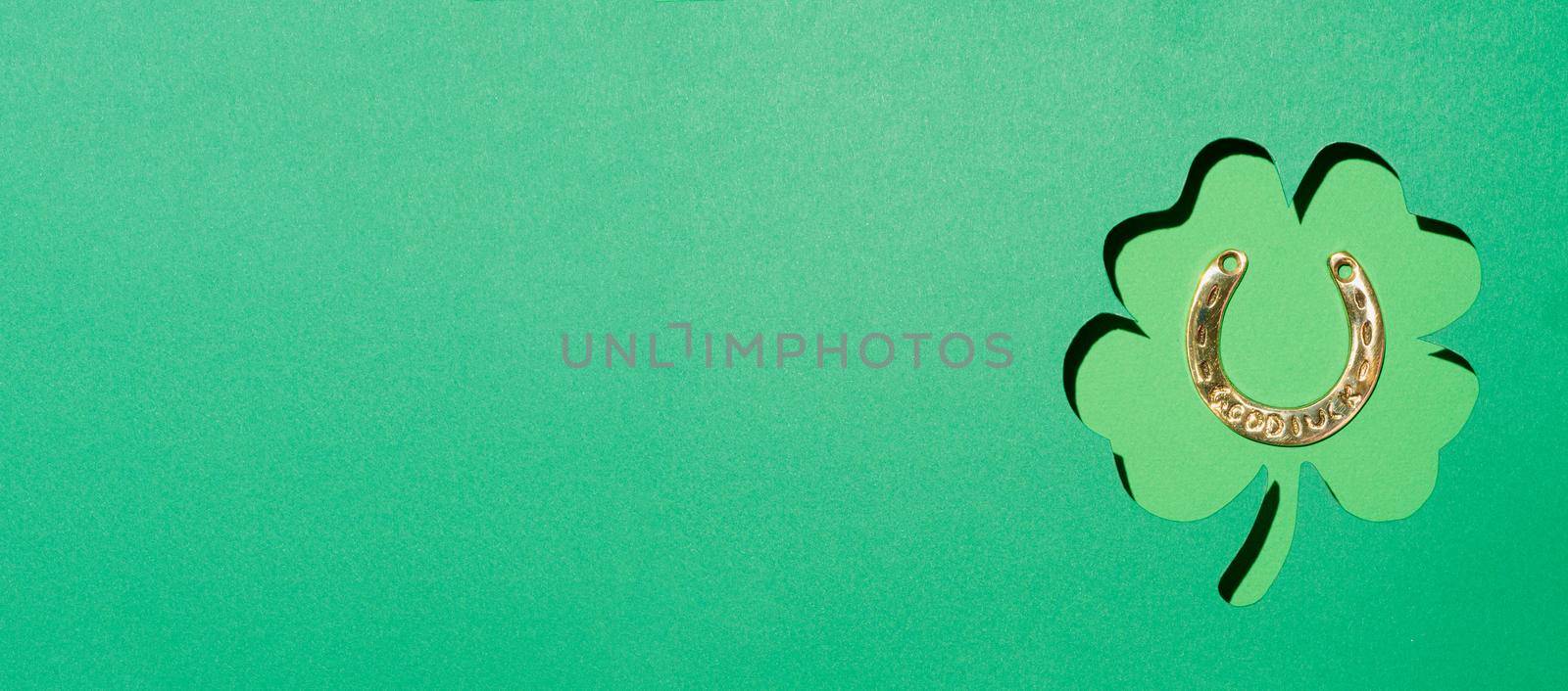 St Patrick's Day concept. Green paper background with cut contour of shamrock leaf and golden horseshoe into leaf shape. Top view or flat lay. Greeting card for Saint Patrick's Day, copy space. Banner