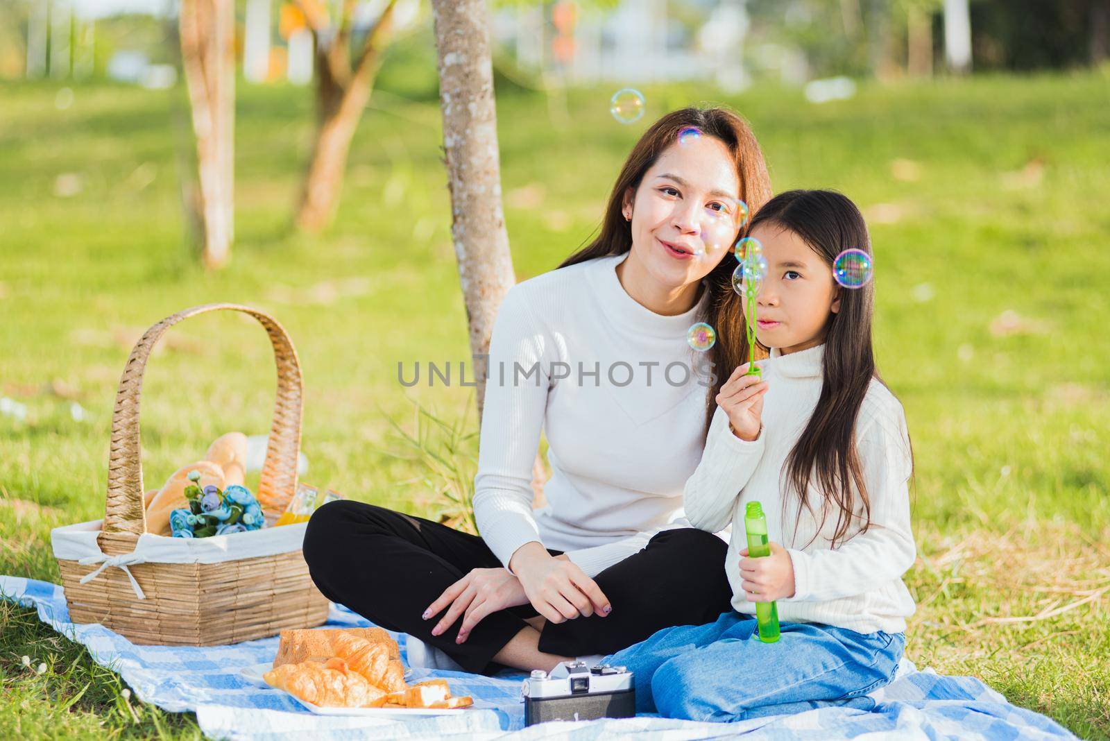 Happy Asian mother and little girl daughter child having fun and enjoying outdoor together sitting on the grass blowing soap bubbles during a picnic in the garden park on a sunny day family time