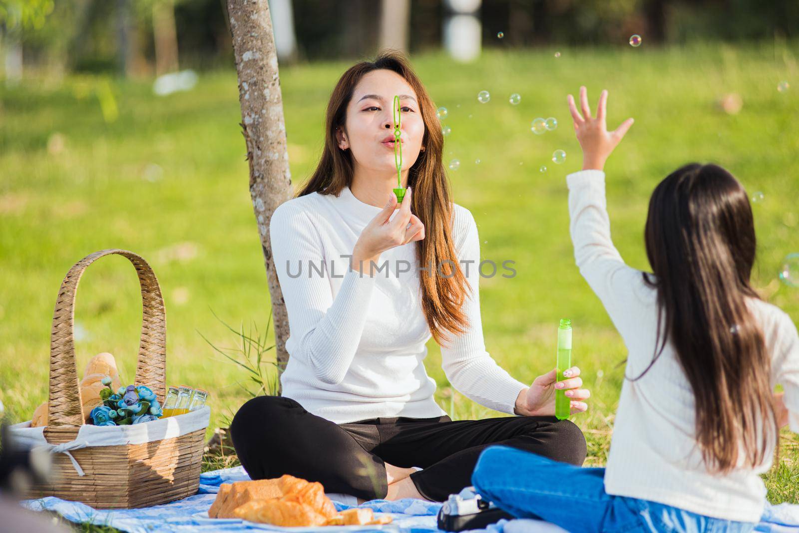 Happy Asian mother and little girl daughter child having fun and enjoying outdoor together sitting on the grass blowing soap bubbles during a picnic in the garden park on a sunny day family time