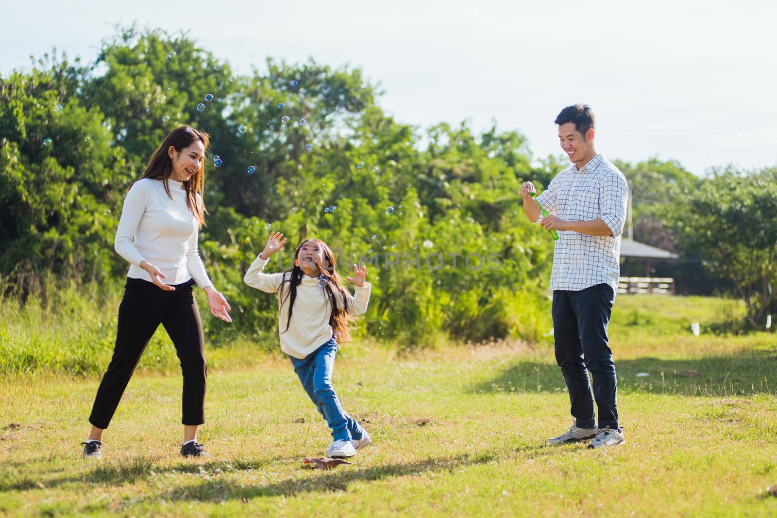 Asian family mother, father and little girl having fun together play blowing soap bubbles in park by Sorapop