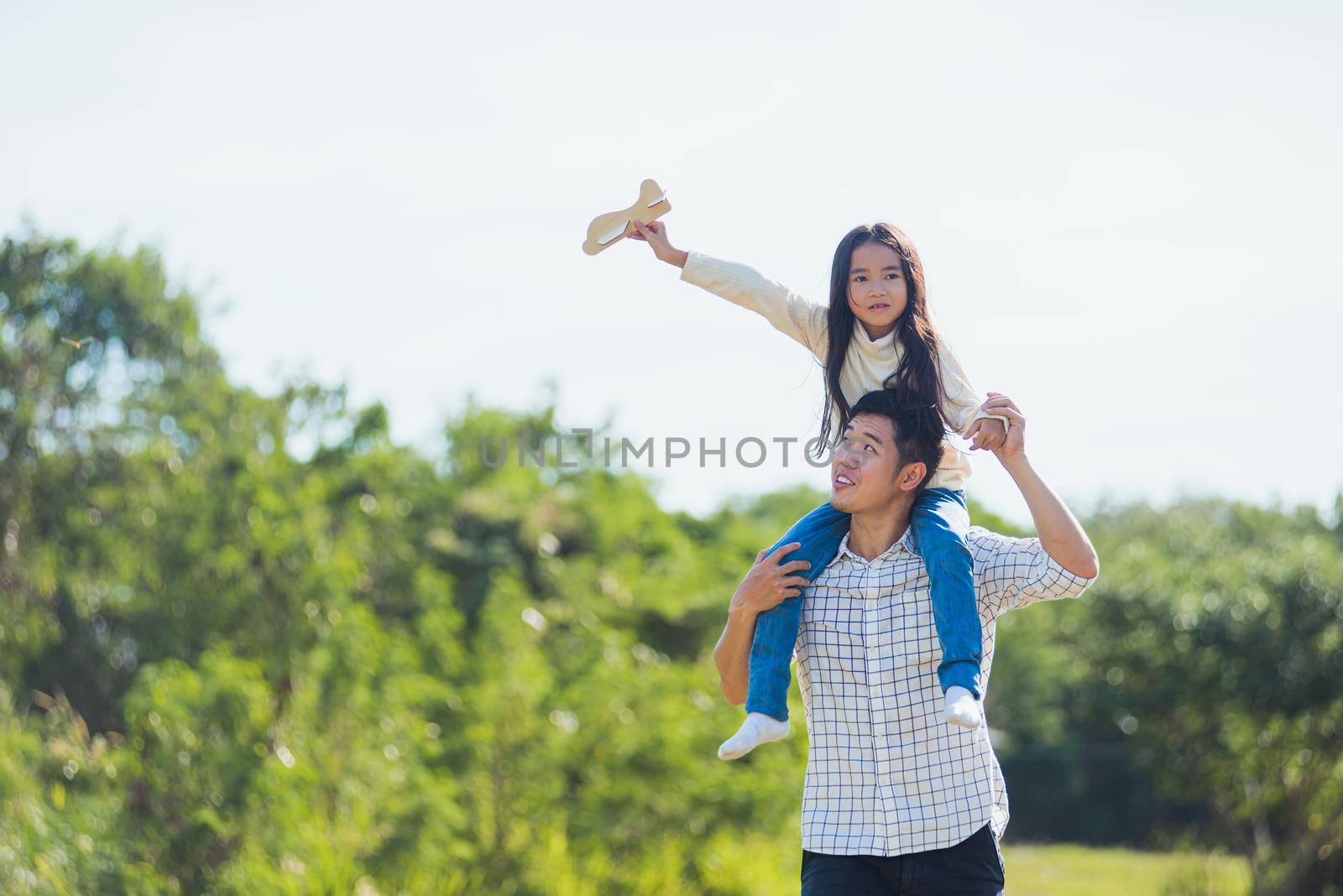 Happy Asian young family father and carrying an excited girl on shoulders having fun and enjoying outdoor lifestyle together playing aircraft toy on sunny summer day, Father's day concept