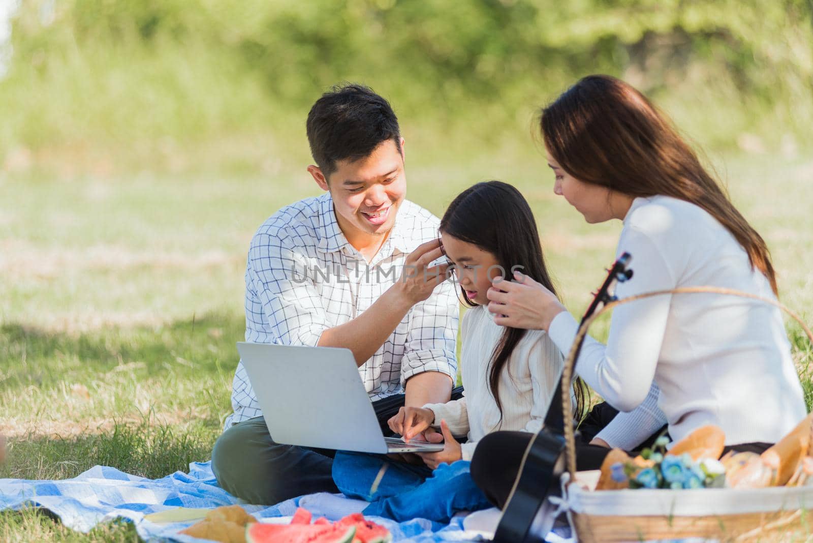 Happy Asian lifestyle young family father, mother and little girl having fun outdoor sitting on picnic blanket using laptop computer technologies while relaxing to open song at garden park in summer