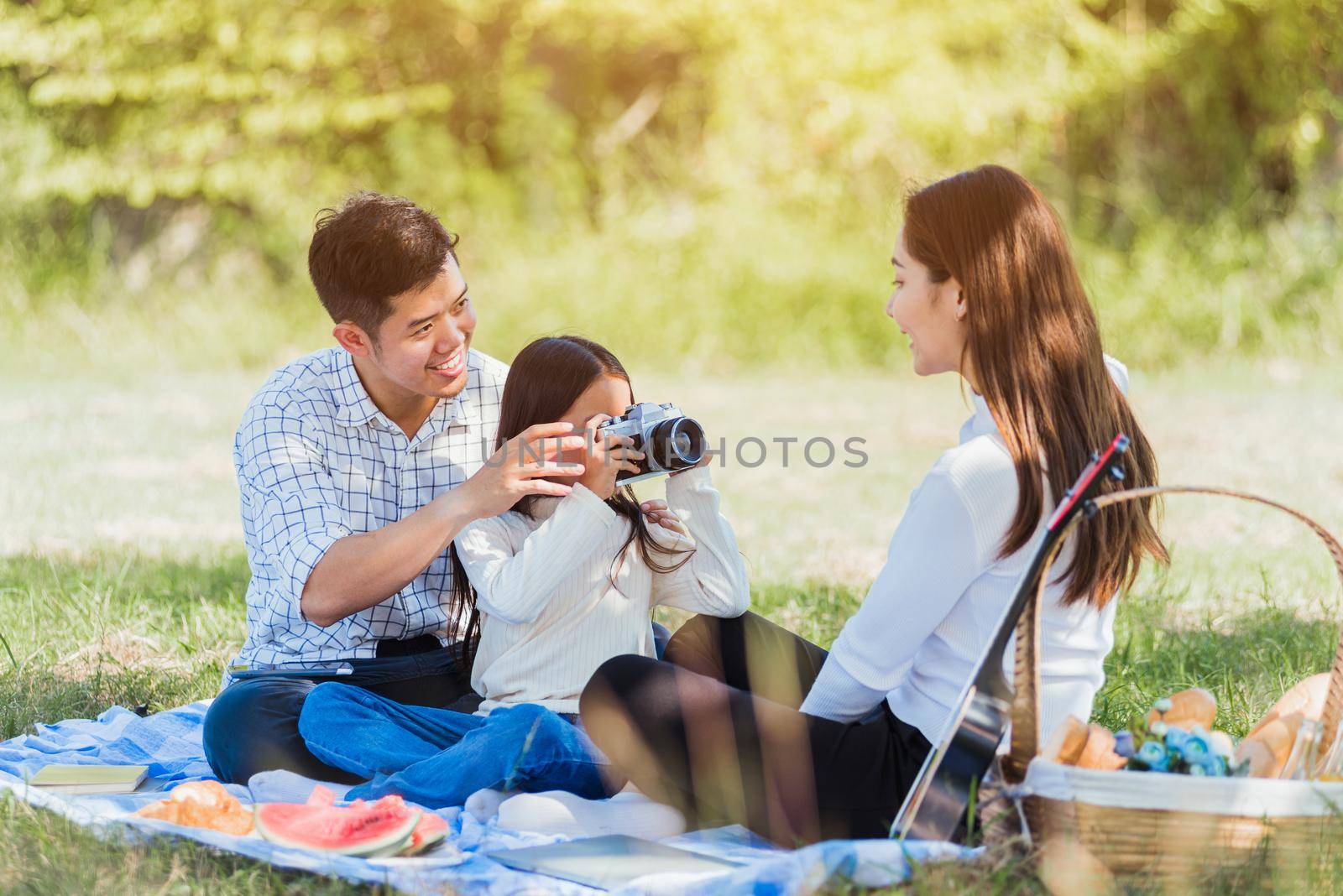 Happy Asian young family father, mother and child little girl having fun and enjoying outdoor picnic together sitting on picnic blanket shooting photos by retro camera in the garden park on sunny day