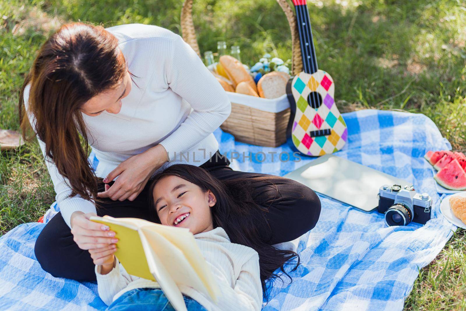 Happy Asian young family mother and child little girl having fun and enjoying outdoor laying on picnic blanket reading book at summer garden spring park, Family relaxation concept