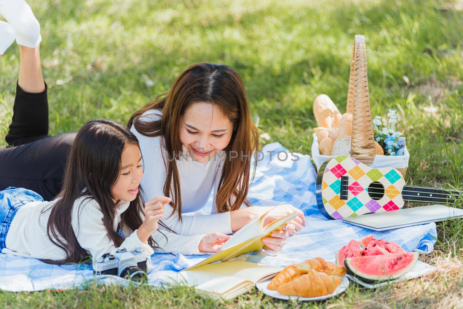 Happy Asian young family mother and child little girl having fun and enjoying outdoor laying on picnic blanket reading book at summer garden spring park, Family relaxation concept