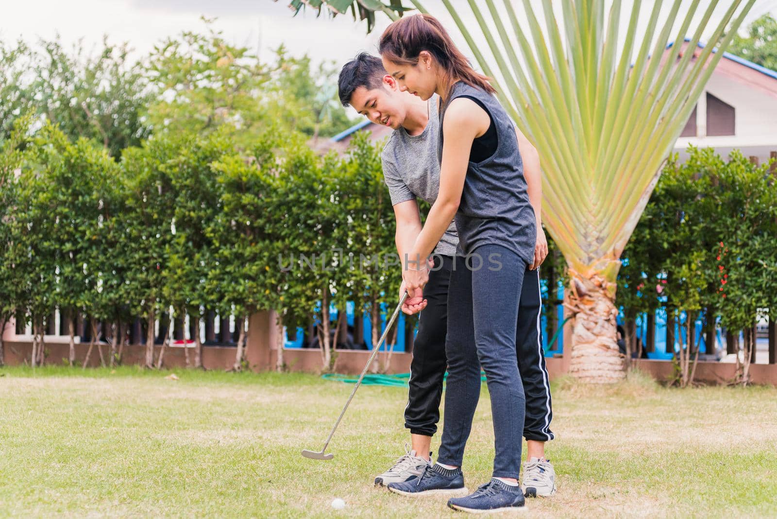 Asian young man support teaching training woman to play perfect golf while standing together in nature a field garden park. Couple trainer giving a lesson on the golf course