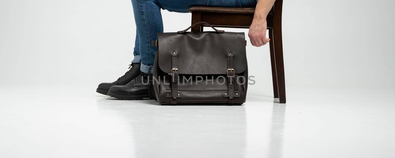 Man in a blue jeans and black boots sits on a chair with a brown men's shoulder leather bag for a documents and laptop on a white floor. Mens leather satchel, messenger bags, handmade briefcase. by vovsht