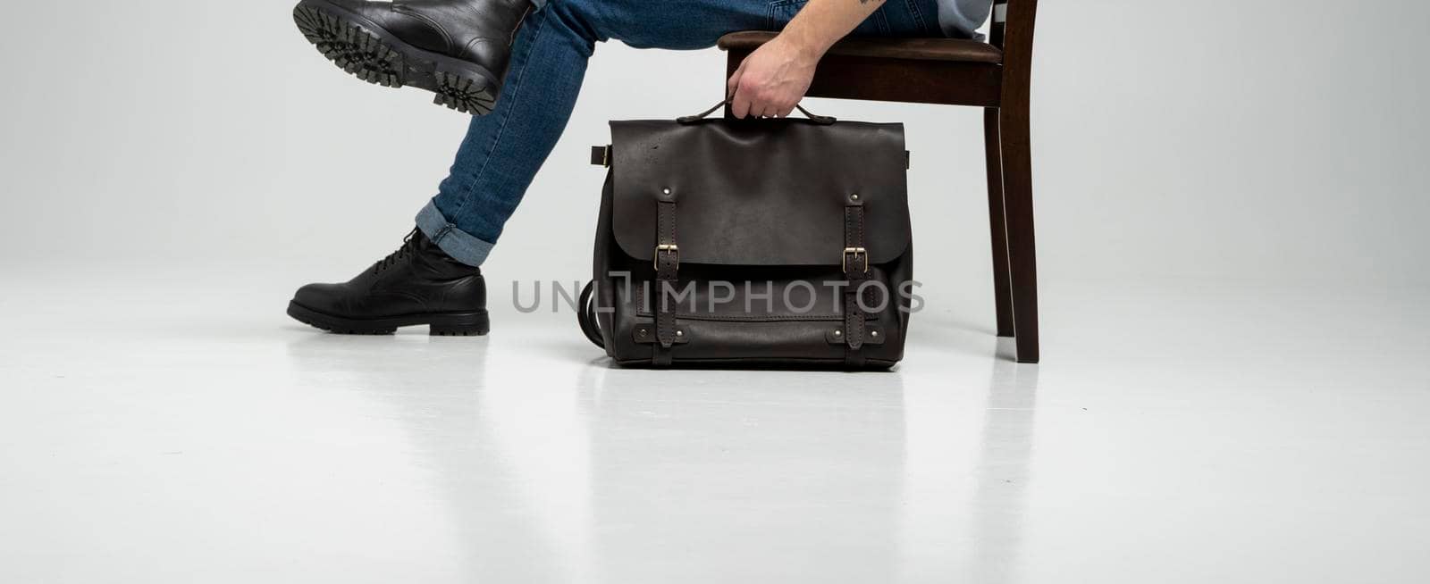 Man in a blue jeans and black boots sits on a chair with a brown men's shoulder leather bag for a documents and laptop on a white floor. Mens leather satchel, messenger bags, handmade briefcase