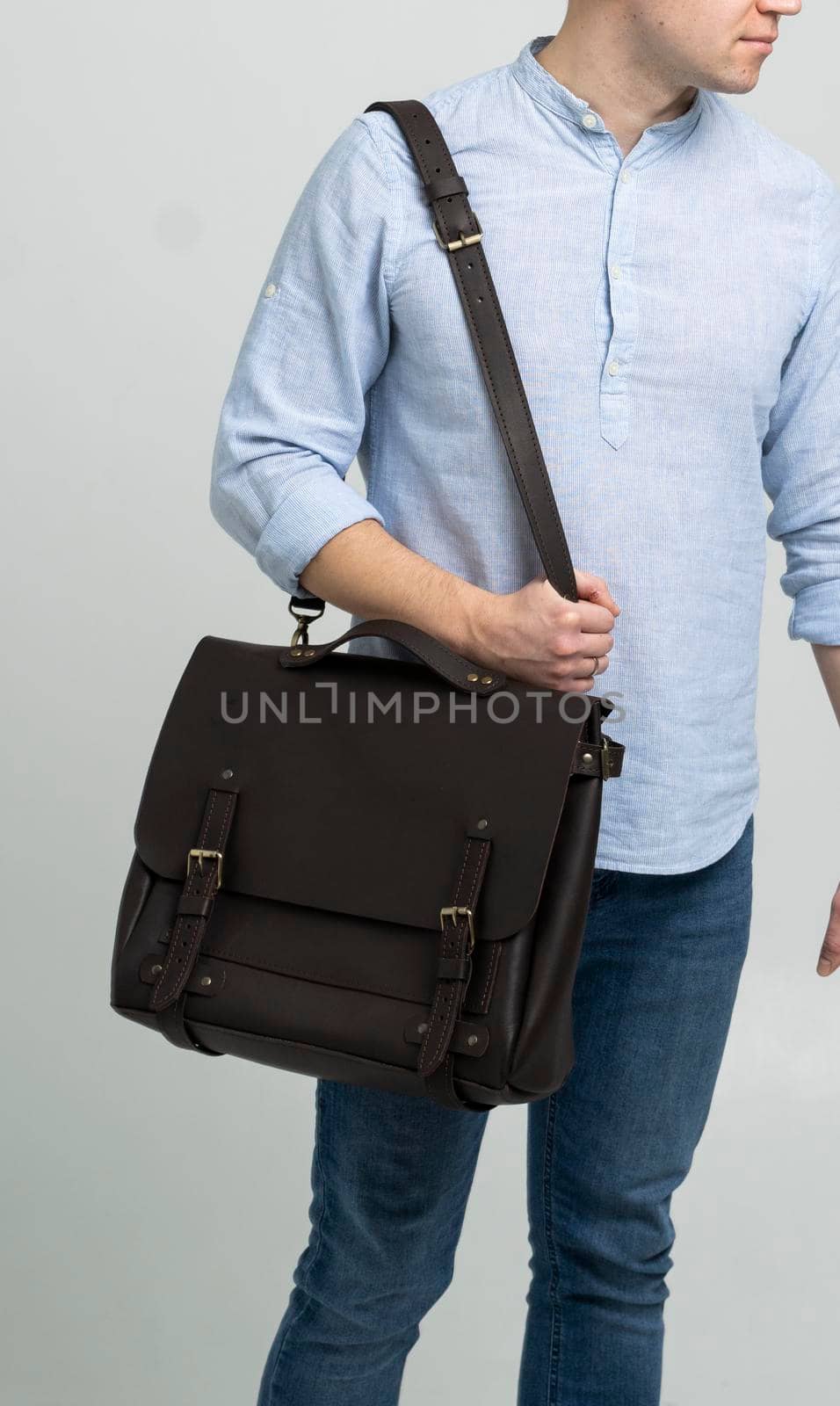 Brown men's shoulder leather bag for a documents and laptop on the shoulders of a man in a blue shirt and jeans with a white background. Satchel, mens leather handmade briefcase