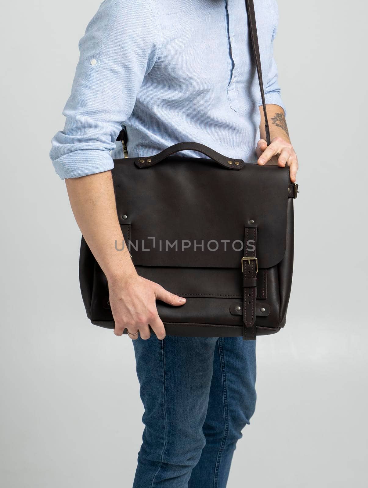 Brown men's shoulder leather bag for a documents and laptop on the shoulders of a man in a blue shirt and jeans with a white background. Satchel, mens leather handmade briefcase
