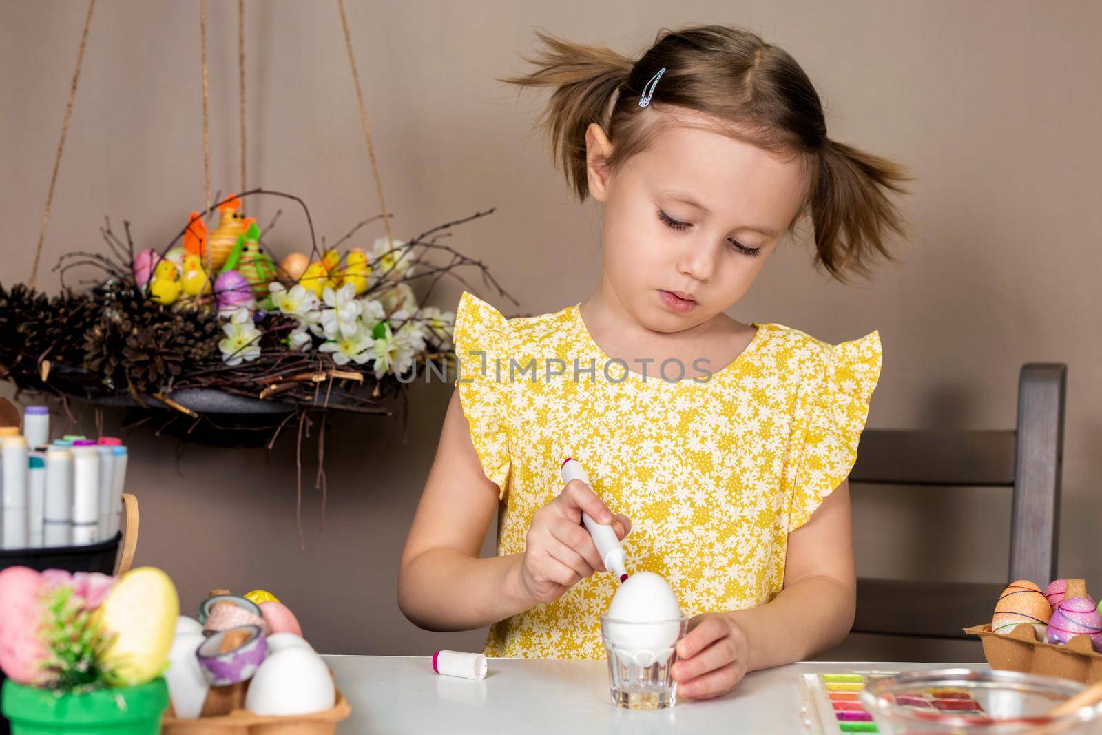 Little adorable girl in yellow dress painting Easter eggs with paintbrush by galinasharapova