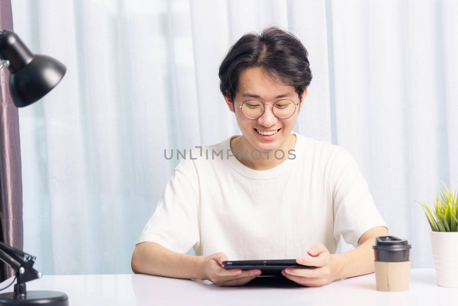 Happy Asian young business handsome man work from home office wear glasses, t-shirt comfortable he smiling and using a black modern smart digital tablet computer to read email or learning on desk