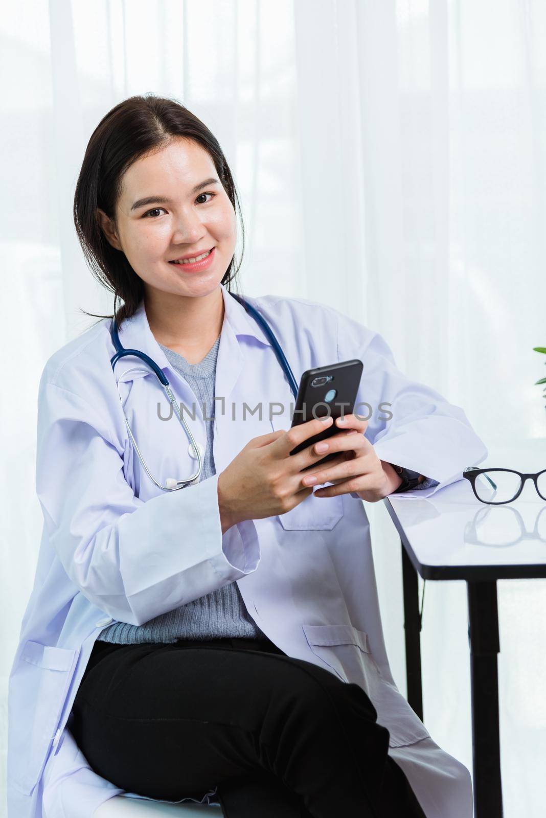 Asian doctor young beautiful woman smiling using working or holding with smart mobile phone and laptop computer at hospital desk office, technology healthcare medical concept