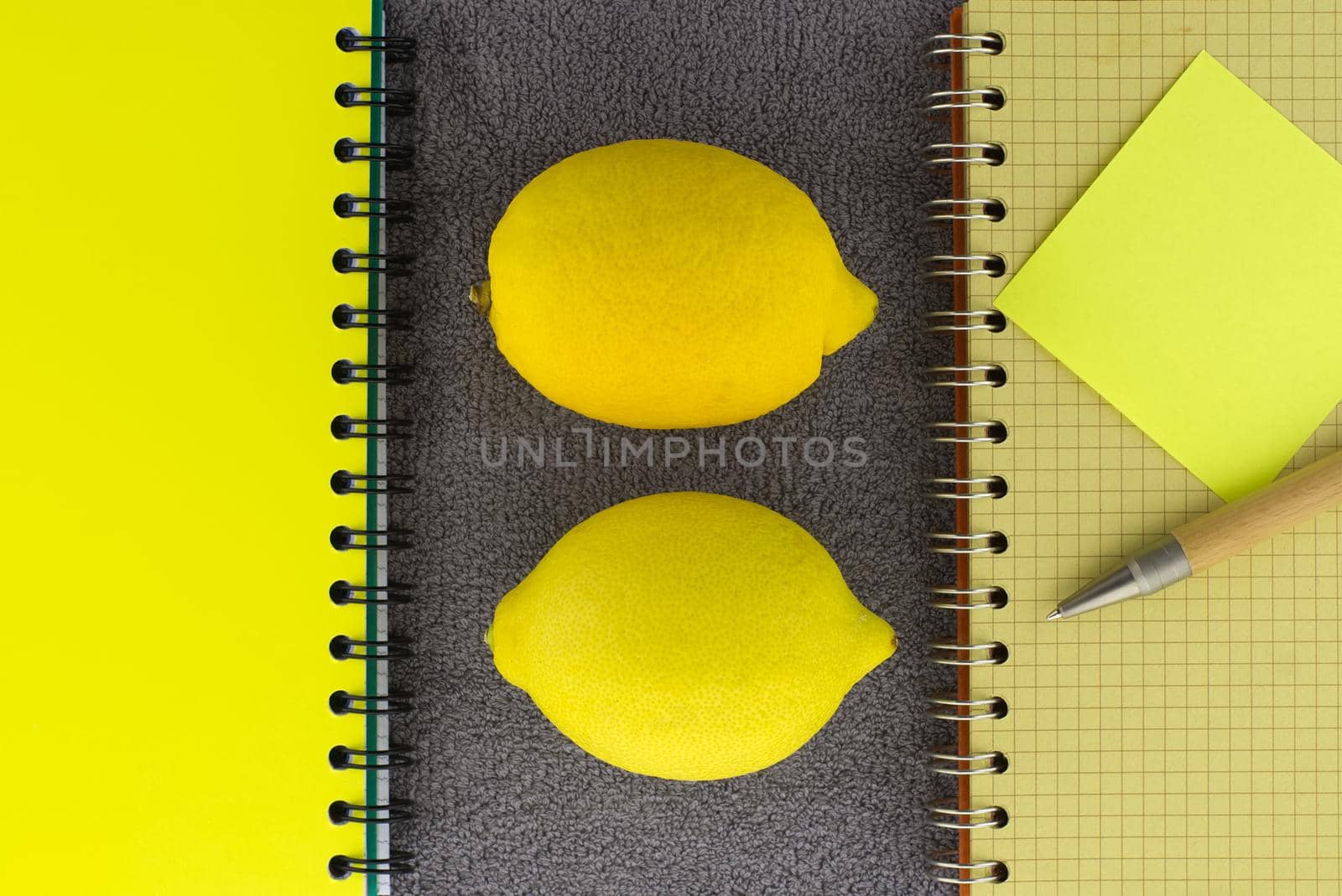 Colorful yellow themed still life with fresh healthy lemons placed to the side of wire bound notepads and pen on a textured grey background viewed as a flat lay from overhead