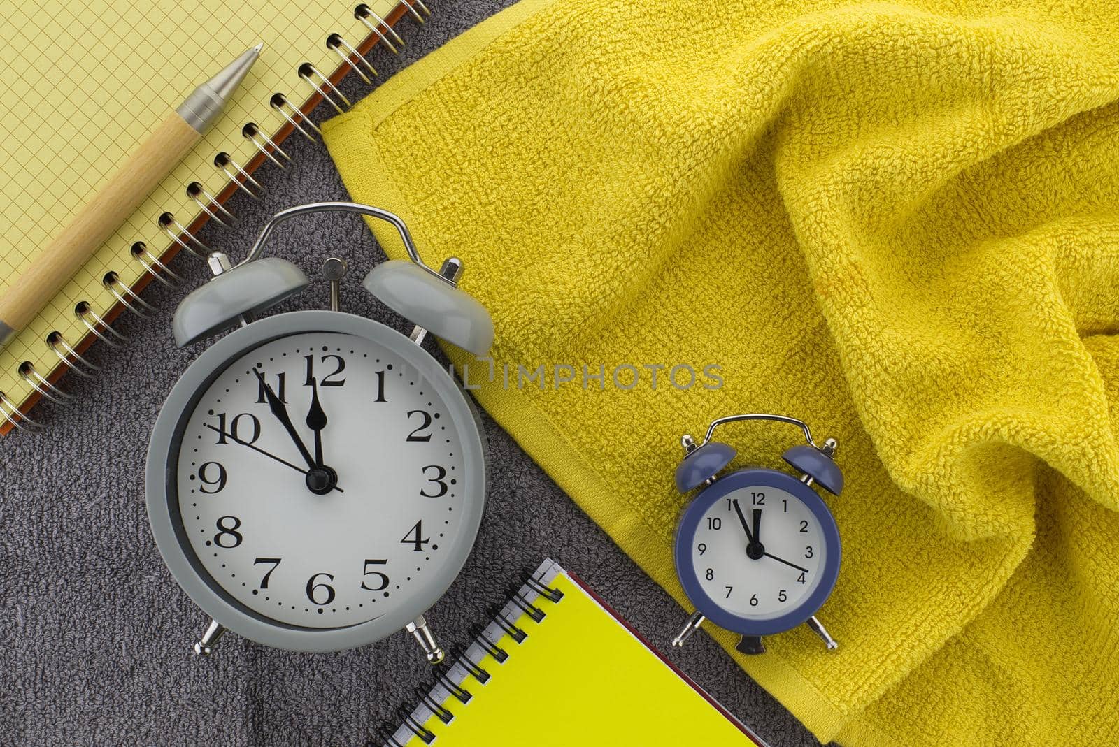 Color trends themed still life with alarm clock placed between of yellow wire bound notepads and pen on a textured grey background viewed as a flat lay from overhead