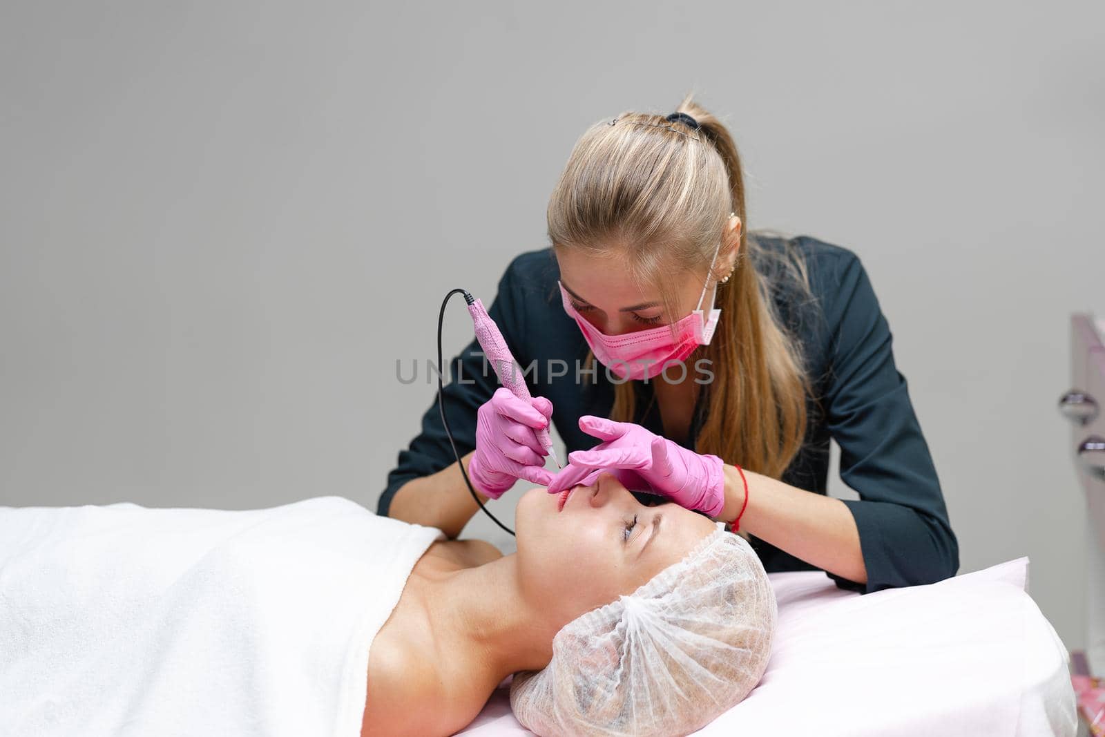 Cosmetology cabinet client lies on couch. Beautician applies permanent ink on lips. Procedure of permanent lip makeup. Free space. Beauty industry