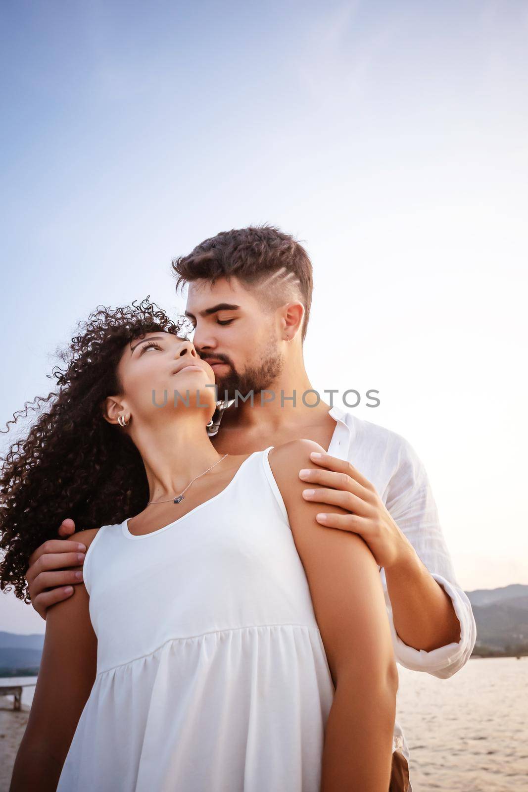 Young macho man in white shirt holding from the shoulders his Hispanic curly beautiful girlfriend. Couple in love looking in the eyes each other outdoor in a sea resort at sunset for valentine day