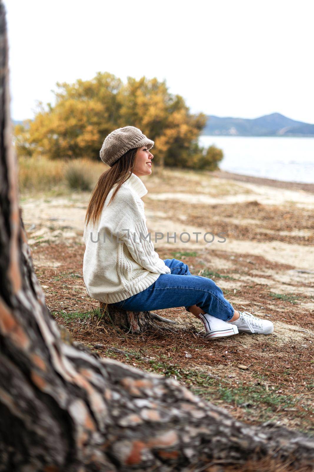 Beautiful Caucasian young woman sitting near the water on a tree trunk in pine forest enjoying the magic winter sea. Happy female person lives the embrace of nature without needing anything else