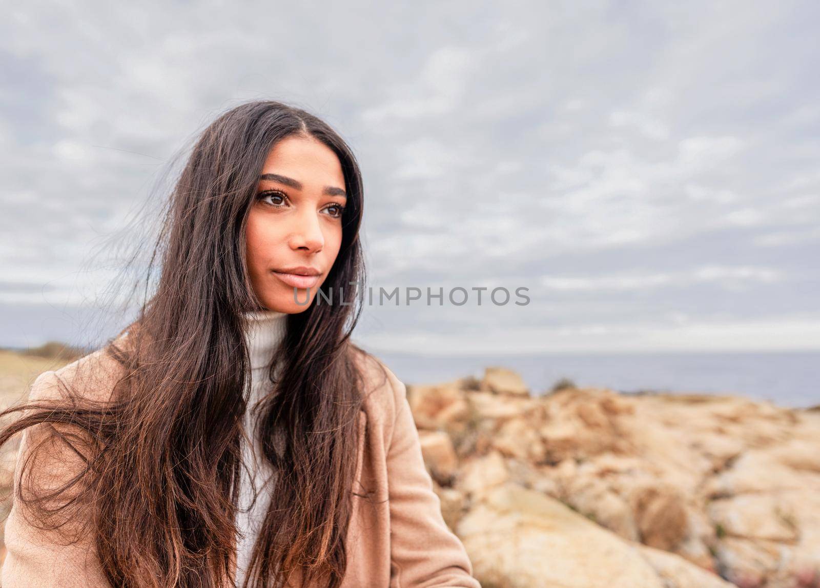 Close up portrait of young beautiful Hispanic woman with long dark hair sitting on sea rocks dreaming looking to the horizon with dramatic grey sky. Fall-winter color mood photography with copy space