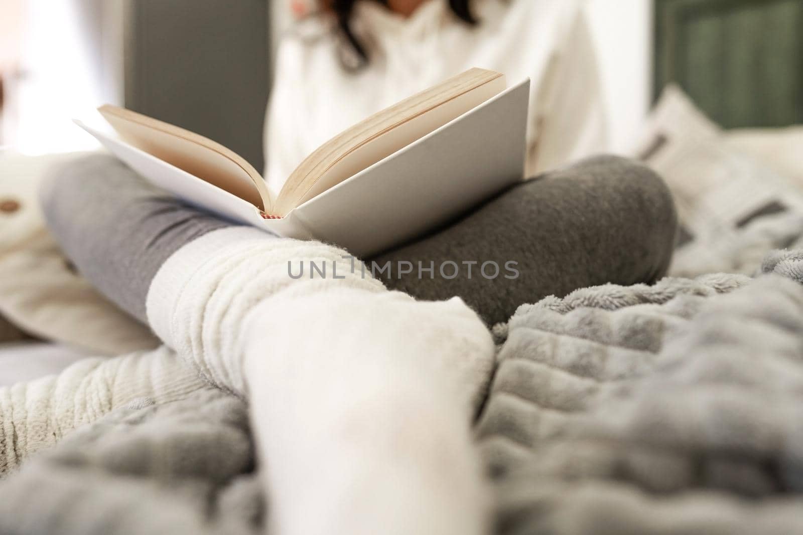 Low angle view of unrecognizable woman sitting on sofa in comfortable clothes relaxing reading a paper book with white wool socks blurred in foreground. Selective focus on the cover