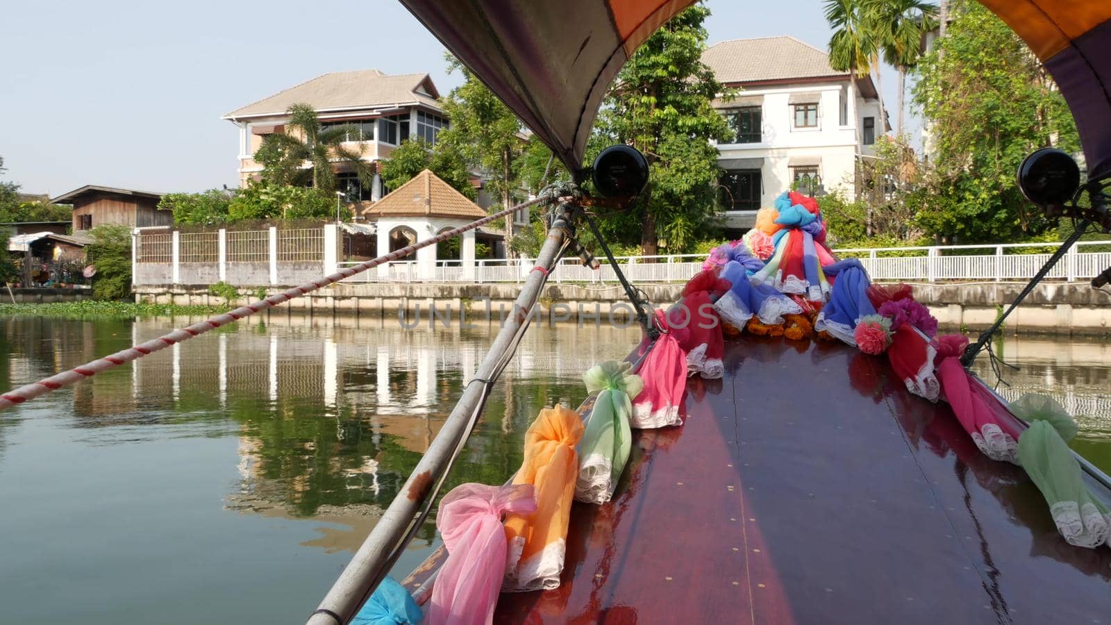 Tourist trip on Asian canal. View of calm channel and residential houses from decorated traditional Thai boat during tourist trip in Bangkok. by DogoraSun