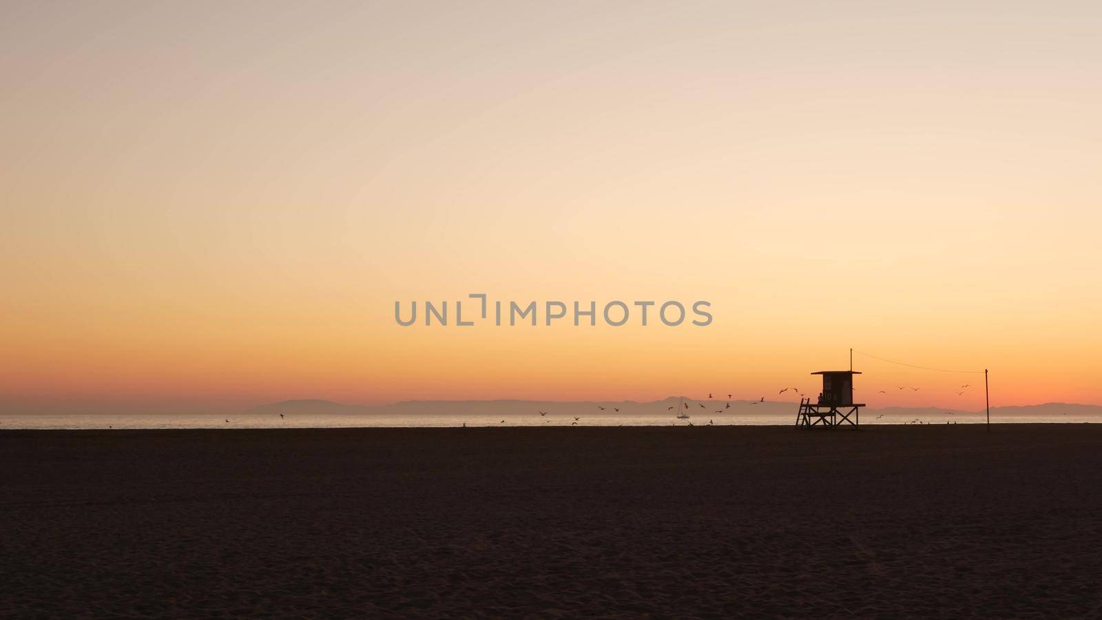 Summertime travel concept. Dark silhouette, iconic retro wooden lifeguard watch tower against sunset orange sky. Contrast watchtower outline, california pacific ocean beach twilight aesthetic, USA.