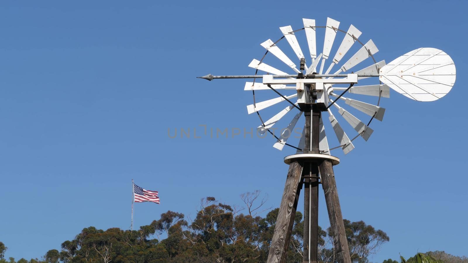 Classic retro windmill, bladed rotor and USA flag against blue sky. Vintage water pump wind turbine, power generator on livestock ranch or agricultural farm. Rural symbol of wild west, rustic suburb.