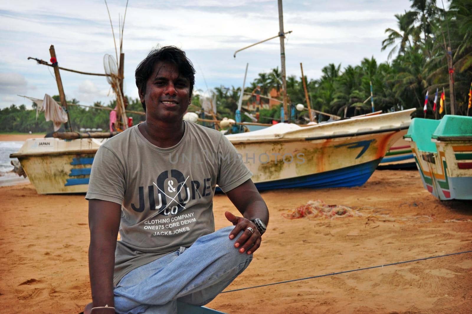 HIKKADUWA, SRI LANKA - 18 DECEMBER 2011 Adult casual ethnic man sitting on remote tropical beach of Sri Lanka smiling at camera. Smiling Indian fisherman on beach