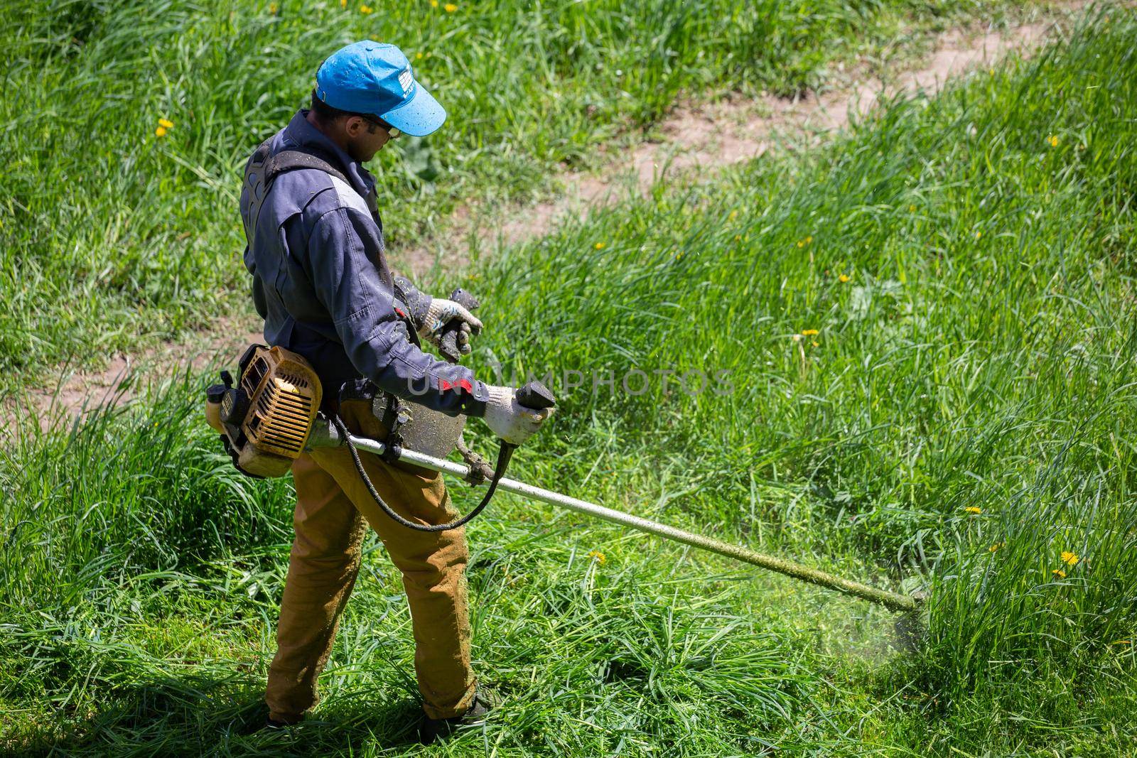 TULA, RUSSIA - MAY 19, 2020: Russian official lawnmower worker man cutting green grass with two-cycle engine string trimmer. Top to down view with selective focus.