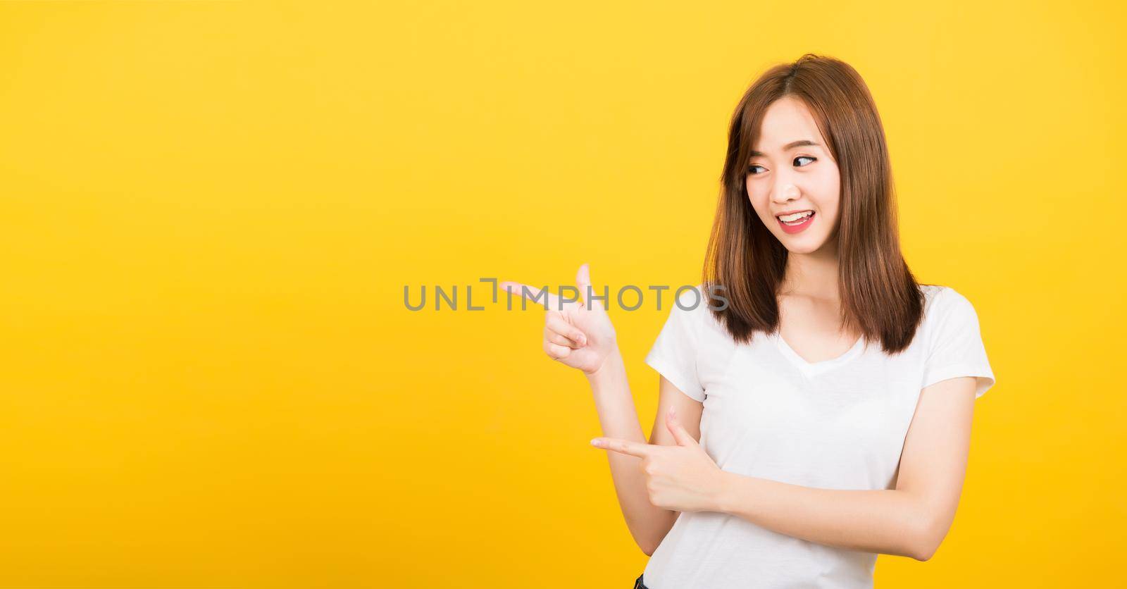 Asian happy portrait beautiful cute young woman teen standing wear t-shirt pointing finger away side looking to side space isolated, studio shot on yellow background with copy space